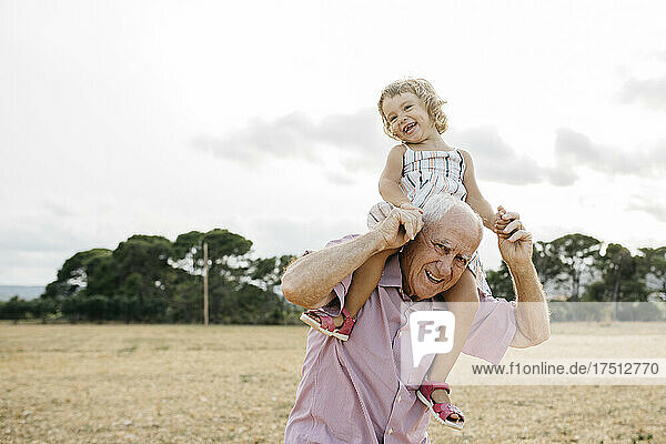 Happy grandfather carrying granddaughter on shoulders while standing against sky