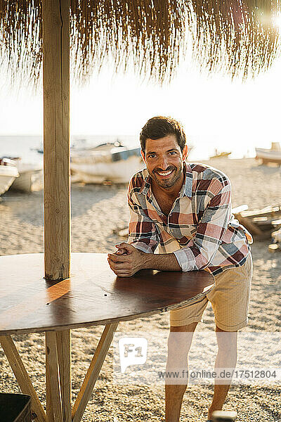 Happy mid adult man standing by table at beach during sunset