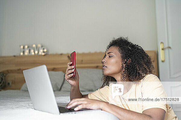 Young woman with curly hair using smart phone and laptop on bed at home