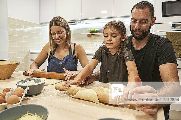 Parents with daughter kneading pizza dough on table in kitchen