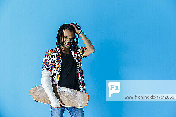 Smiling man with fractured arm holding skateboard against blue background