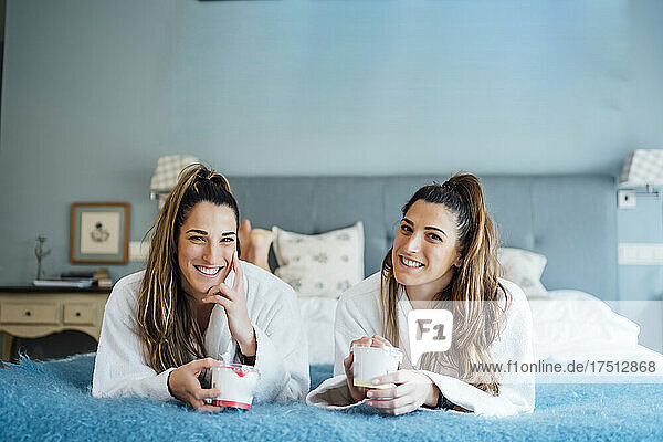 Smiling town sisters holding container while lying on bed in hotel room