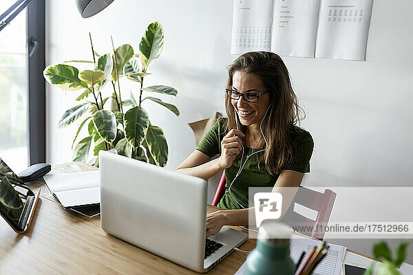 Cheerful female entrepreneur wearing headphones working over laptop on desk at home