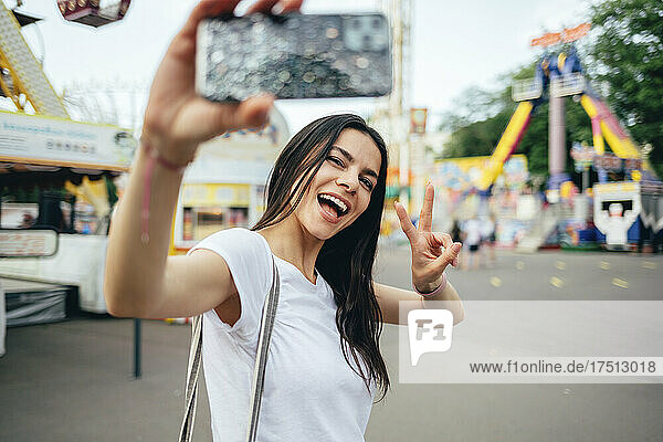 Glückliche junge Frau gestikuliert  während sie im Vergnügungspark ein Selfie macht