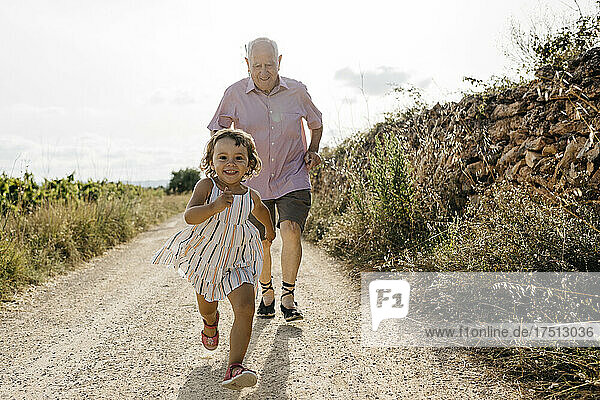 Grandfather running behind playful granddaughter on dirt road against sky