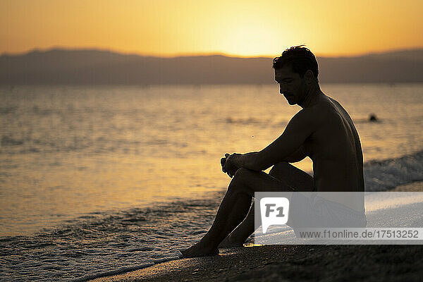 Shirtless thoughtful mid adult man sitting at shore against sky during sunset