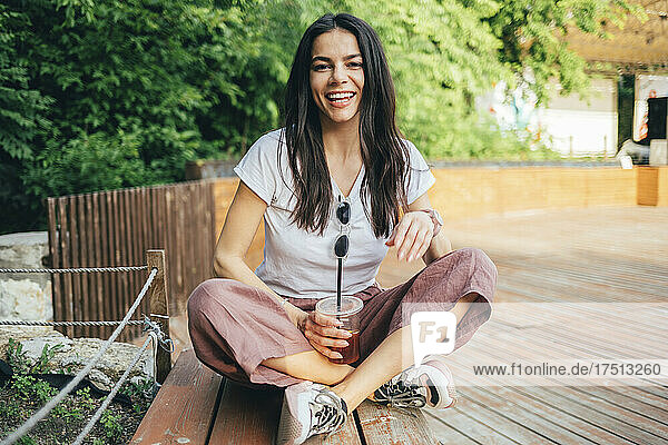 Young cheerful woman holding coffee while sitting on bench in park