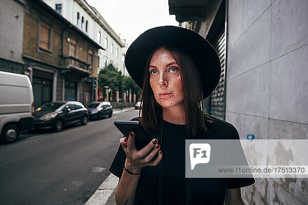 Young woman wearing hat holding smart phone while looking away in city