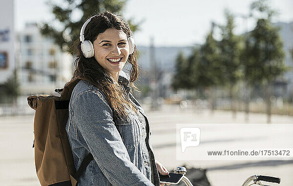 Smiling female student listening music through headphones standing in city on sunny day