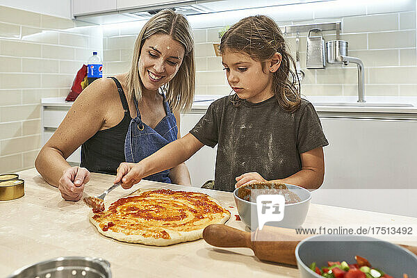 Mother looking at daughter applying sauce on pizza dough in kitchen