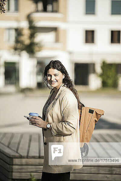 Junge Frau trägt Rucksack  hält Smartphone in der Hand und trinkt  während sie in der Stadt steht