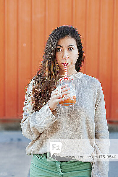 Beautiful woman drinking juice while standing against orange wall