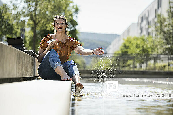 Cheerful woman holding smart phone playing with pond in park during sunny day
