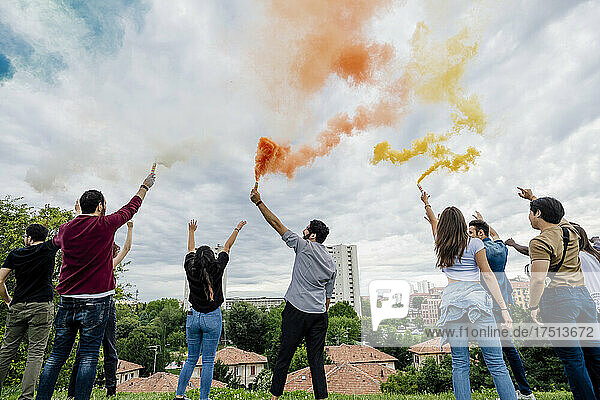 Male and female friends playing with smoke bombs against cloudy sky in park