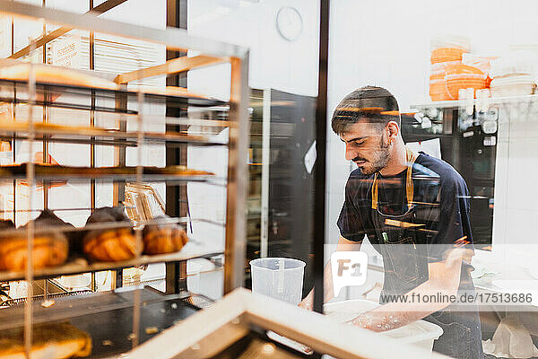 Young baker working at bakery seen through glass window