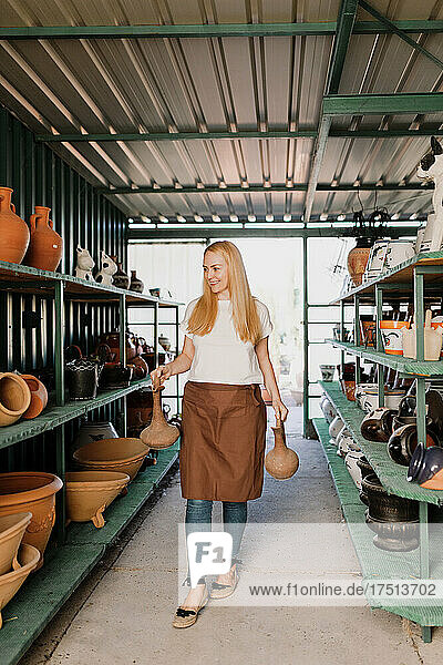 Smiling female owner holding potteries while walking amidst shelves in workshop