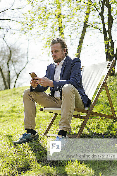 Mature businessman text messaging through mobile phone while sitting on chair at park