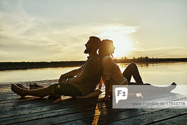 Couple sitting back to back on jetty at a lake at sunset