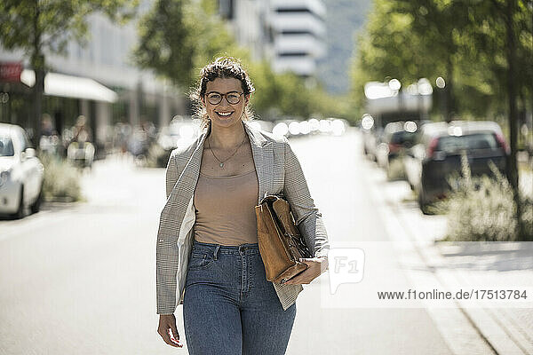 Smiling young woman carrying bag while walking on road during sunny day