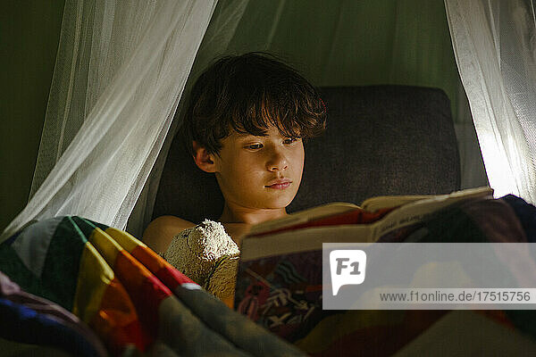 a young boy sits in chair with stuffed toy reading book at bedtime
