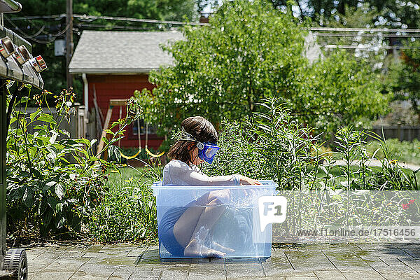 Girl in swimsuit and goggles sits in small plastic tub of water