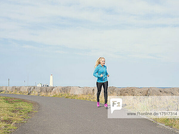 Sporty female jogging on path near sea on sunny day