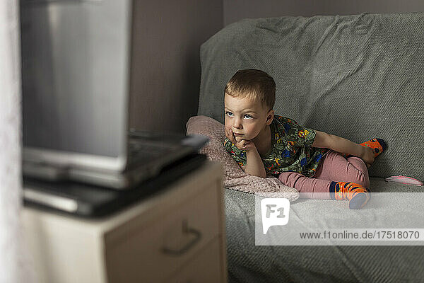 Small blonde boy lying down on sofa and watching cartoons on lap
