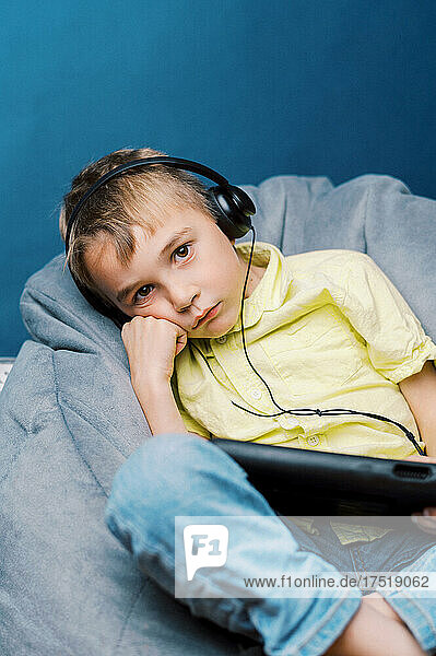 Little sad boy on tablet with headphones on blue background