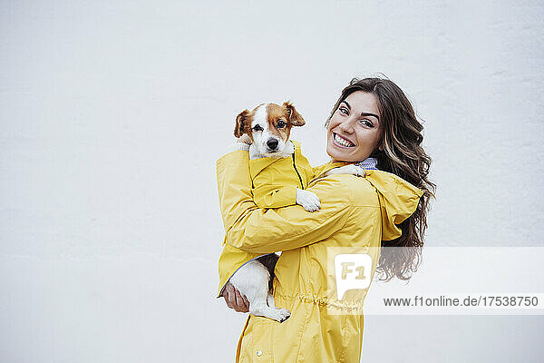 Smiling young woman embracing dog by white wall