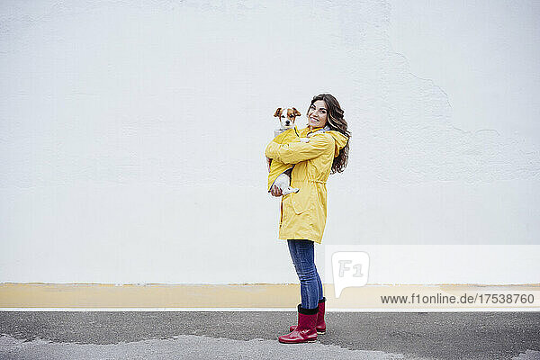 Smiling young woman wearing raincoat carrying dog on road