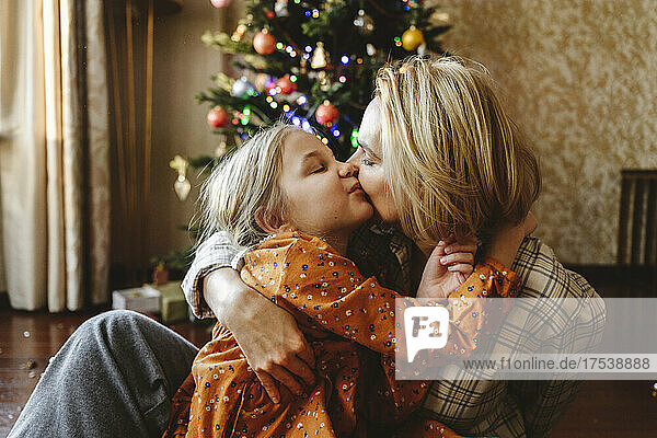 Mother kissing daughter in front of christmas tree at home