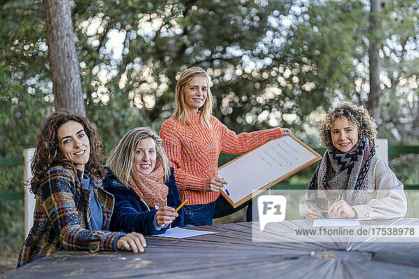 Happy woman holding whiteboard with friends sitting at table in park