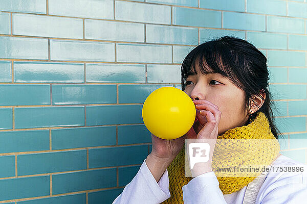 Woman blowing yellow balloon by turquoise brick wall