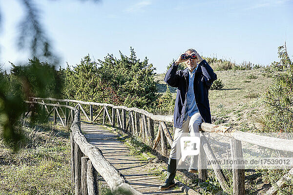 Man looking through binoculars sitting on railing at wooden bridge