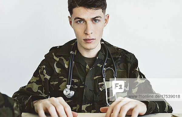 Army doctor with stethoscope sitting in front of white wall