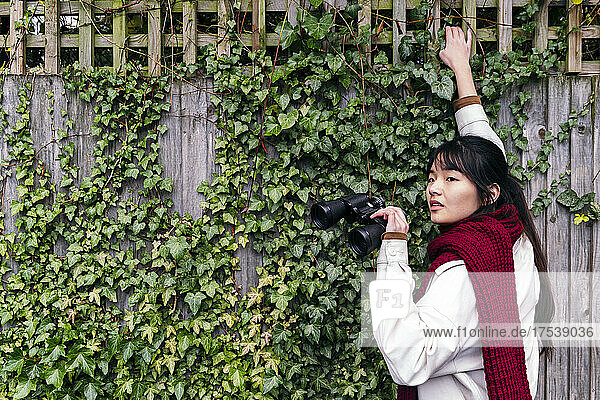 Young woman with binoculars standing near ivy wall