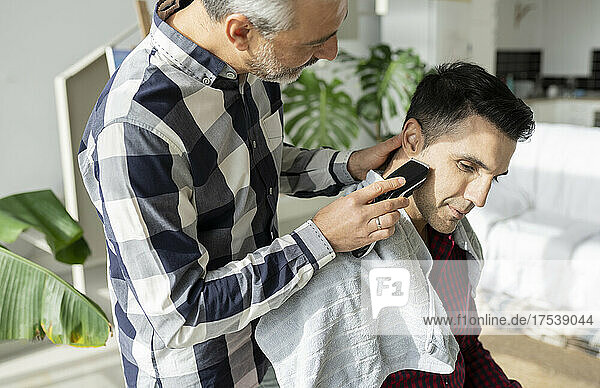 Gay man cutting husband's beard with electric razor at home