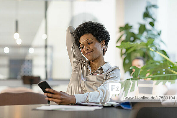 Lächelnde Geschäftsfrau mit der Hand hinter dem Kopf  die im Büro ihr Smartphone benutzt