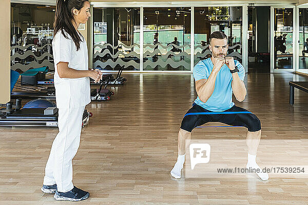 Fitness instructor looking at athlete exercising with resistance band in gym