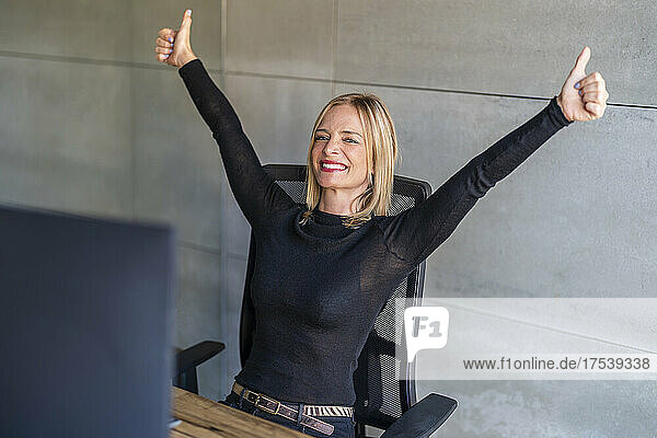 Happy businesswoman showing thumps up sitting on chair in front of gray wall at office