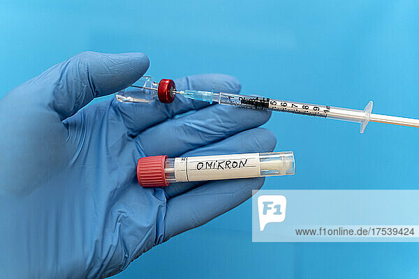 Doctor with protective glove holding swab tube with Omicron and syringe against blue background