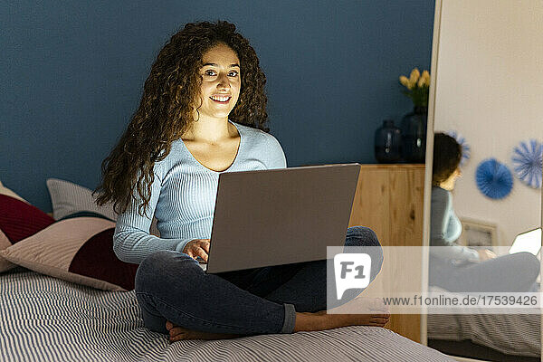 Smiling woman sitting on bed with laptop at home