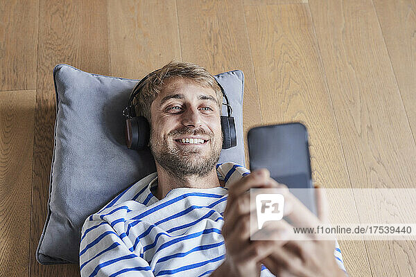 Smiling man with headphones using smart phone lying on hardwood floor