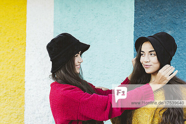 Smiling woman adjusting bucket hat of twin sister by colorful wall