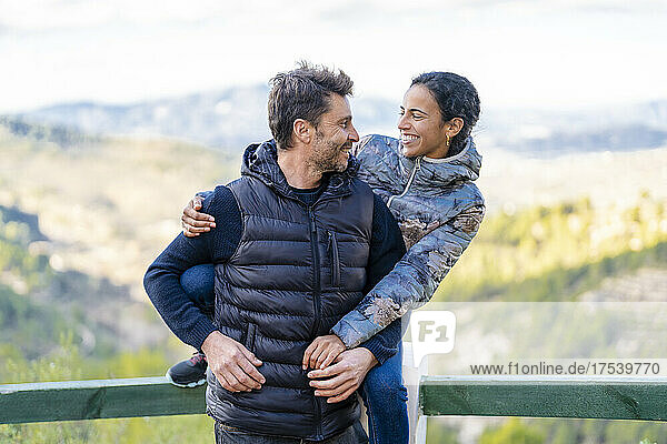 Smiling woman sitting on railing hugging man from behind at park