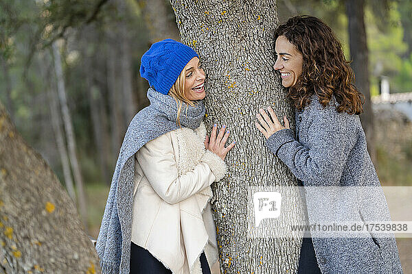 Cheerful friends looking at each other leaning on tree trunk