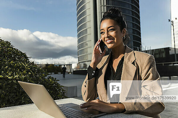 Lächelnde Geschäftsfrau mit Laptop  die auf dem Smartphone spricht