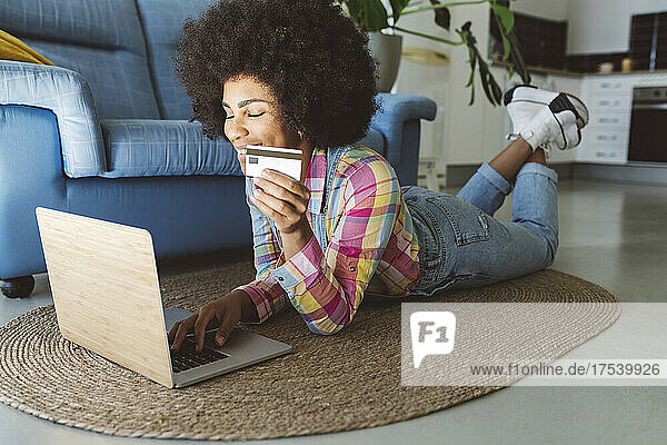 Smiling woman shopping online using laptop and credit card lying on carpet at home