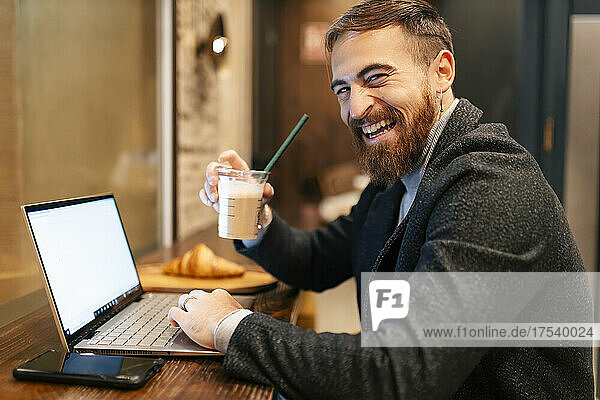 Cheerful businessman holding coffee cup sitting with laptop and smart phone at restaurant