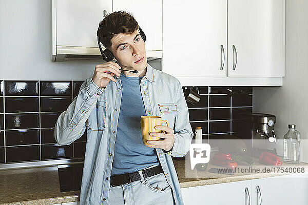 Junger Mann mit Headset und Kaffeetasse lehnt zu Hause auf der Küchentheke